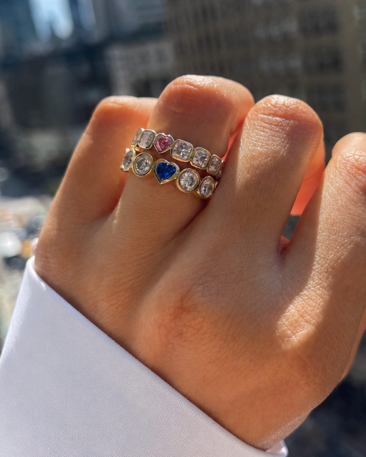 Two stacked Hidden Gem rings with pink and blue sapphire hearts photographed in soft daylight.